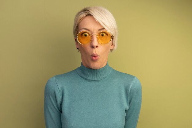 Surprised young blonde woman wearing sunglasses looking at front pursing lips isolated on olive green wall