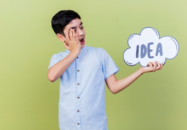 Surprised young boy holding and looking at idea bubble keeping hand on face isolated on olive green wall