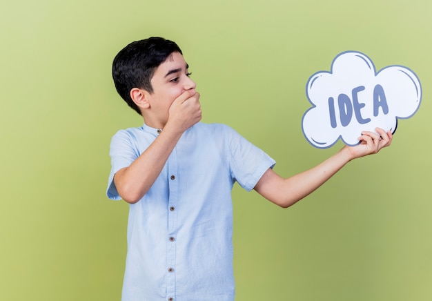 Surprised young boy holding and looking at idea bubble keeping hand on mouth isolated on olive green wall