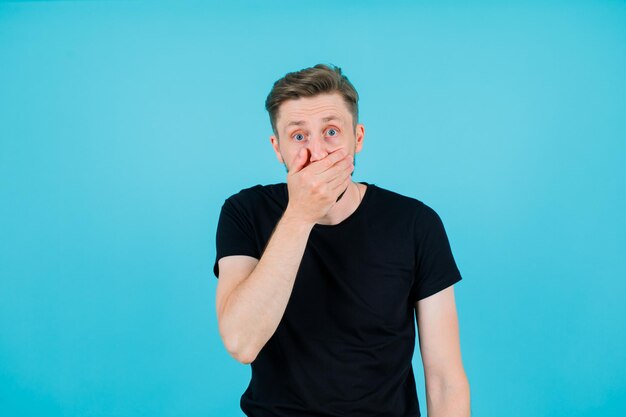 Surprised young boy is putting hand on mouth on blue background