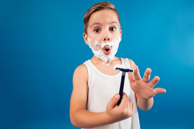 Surprised young boy in shaving foam like man holding razor