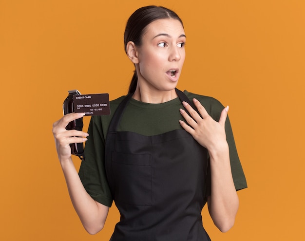 Surprised young brunette barber girl in uniform holds hair clippers and credit card puts hand on chest looking at side on orange
