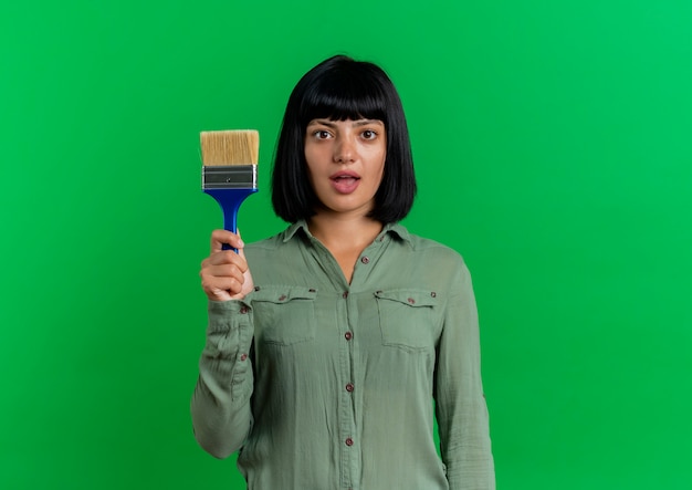 Free photo surprised young brunette caucasian girl holds paint brush isolated on green background with copy space