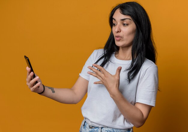 Удивленная молодая кавказская девушка брюнетка смотрит и указывает на телефон с рукой, изолированной на оранжевой стене