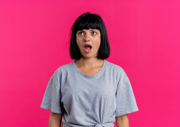 Surprised young brunette caucasian girl looks up isolated on pink background with copy space