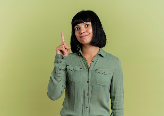 Surprised young brunette caucasian girl points up isolated on olive green background with copy space