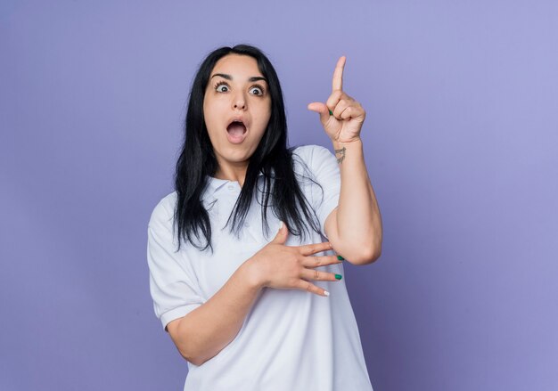 Surprised young brunette caucasian girl points up and looks forward isolated on purple wall