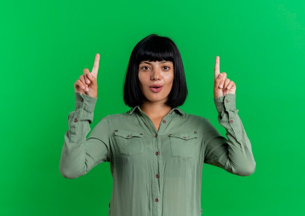 Удивленная молодая брюнетка кавказская девушка показывает вверх двумя руками, изолированными на зеленом фоне с копией пространства