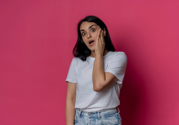 Surprised young brunette caucasian girl puts hand on face looking isolated on pink wall