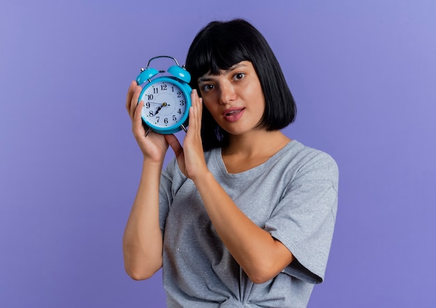 Surprised young brunette caucasian woman holds alarm clock isolated on purple background with copy space