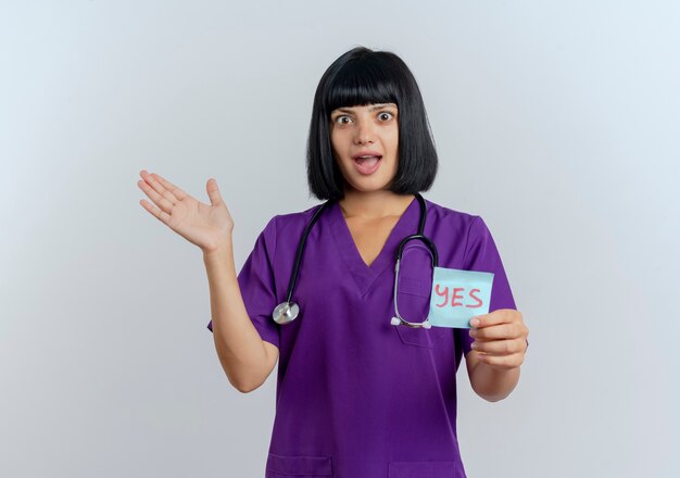 Surprised young brunette female doctor in uniform with stethoscope holds yes note