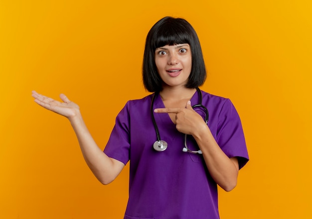 Surprised young brunette female doctor in uniform with stethoscope points at empty hand isolated on orange background with copy space