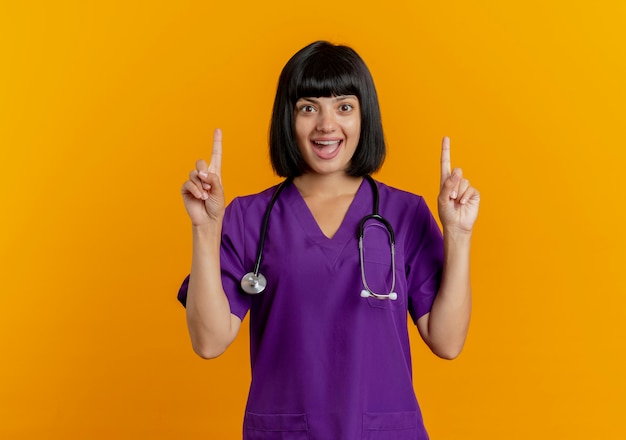 Удивленная молодая брюнетка женщина-врач в униформе со стетоскопом указывает вверх двумя руками