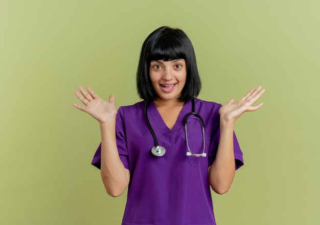 Foto gratuita il giovane medico femminile del brunette sorpreso in uniforme con lo stetoscopio sta con le mani aperte