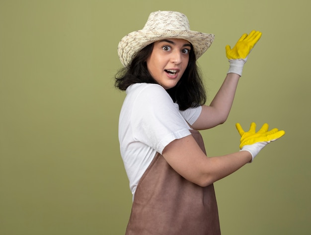 園芸帽子と手袋を身に着けている制服を着た驚いた若いブルネットの女性の庭師は、オリーブの緑の壁に隔離された手で後ろを指します