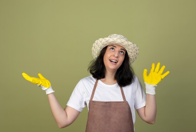 Surprised young brunette female gardener in uniform wearing gardening hat and gloves stands with raised hands looking up isolated on olive green wall