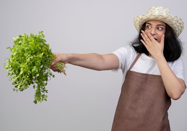 Удивленная молодая брюнетка женщина-садовник в униформе в садовой шляпе кладет руку на рот и держит кориандр, изолированный на белой стене
