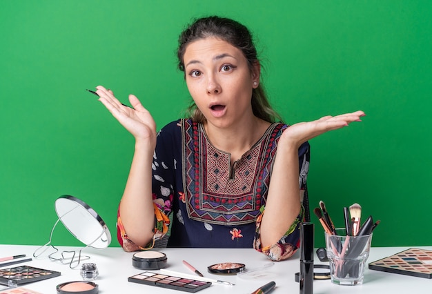 Sorpresa giovane ragazza bruna seduta al tavolo con strumenti per il trucco che tiene le mani aperte tenendo l'eyeliner