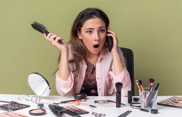 Surprised young brunette girl sitting at table with makeup tools talking on phone and holding comb isolated on olive green wall with copy space