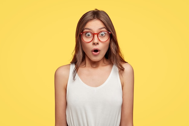 Бесплатное фото Удивленная молодая брюнетка в очках позирует на фоне желтой стены