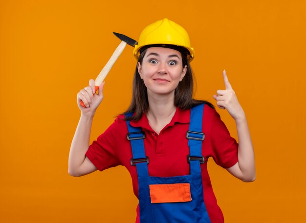 Surprised young builder girl holds hammer and points up on isolated orange background