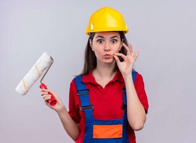 Surprised young builder girl holds paint roller and zipps mouth shut on isolated white background