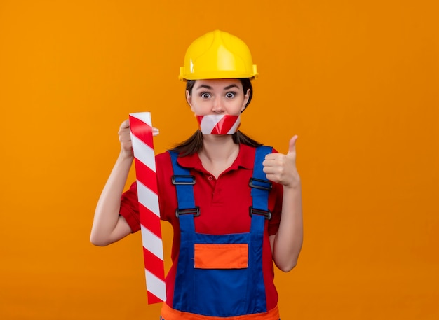 Удивленная молодая девушка-строитель, заклеенная предупреждающей лентой, держит ленту и показывает палец вверх на изолированном оранжевом фоне с копией пространства