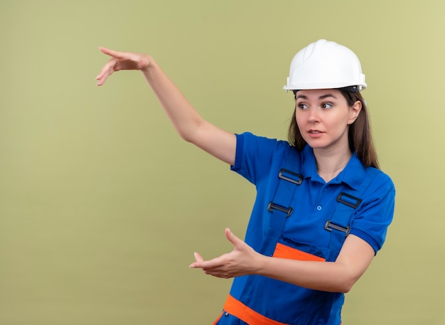 Surprised young builder girl with white safety helmet and blue uniform pretends to hold something with both hands on isolated green background with copy space