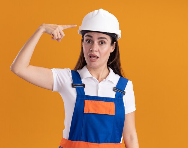 Surprised young builder woman in uniform points at herself isolated on orange wall