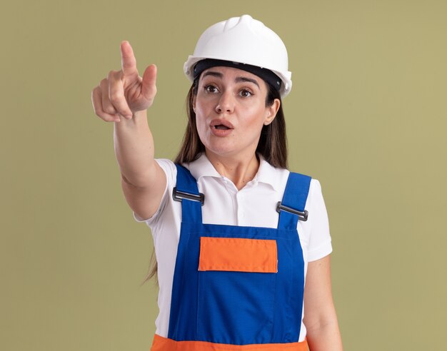 Surprised young builder woman in uniform points at side isolated on olive green wall