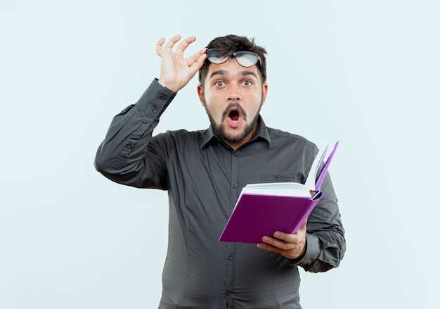 Free photo surprised young businessman wearing glasses holding book and grabbed glasses isolated on white