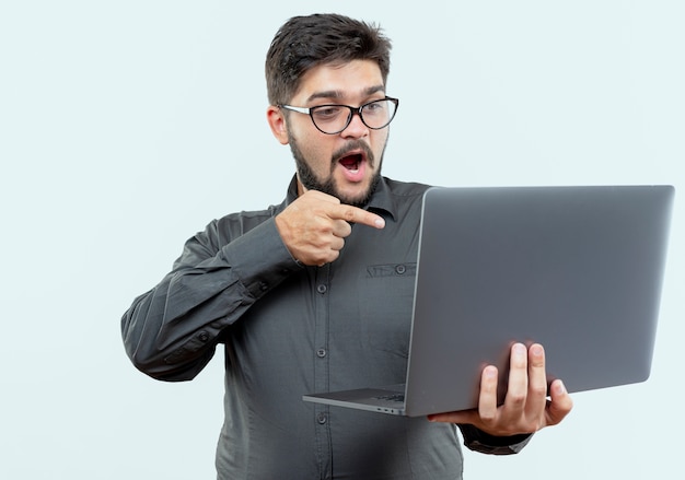 Удивленный молодой бизнесмен в очках держит и указывает на ноутбук, изолированный на белом