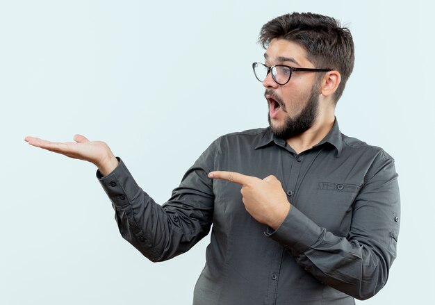 Surprised young businessman wearing glasses pretending holding and points at something isolated on white