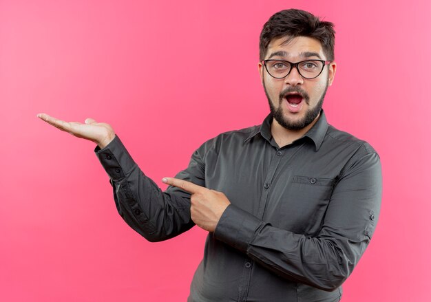 Surprised young businessman wearing glasses pretending holding something isolated on pink