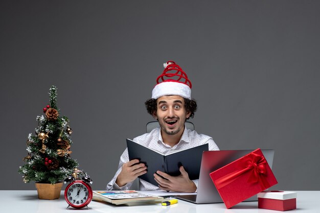 Surprised young businessman with funny santa claus hat checking information in documents in the office on dark background