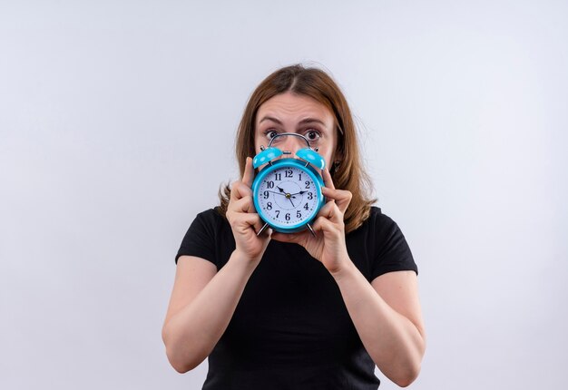 Surprised young casual woman holding and hiding behind alarm clock on isolated white wall with copy space