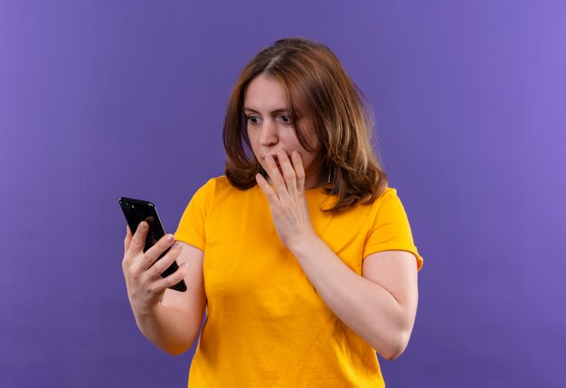 Surprised young casual woman holding mobile phone and looking at it with hand on mouth on isolated purple wall with copy space