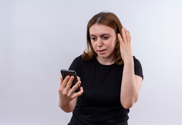 Surprised young casual woman holding mobile phone and looking at it with hand near head on isolated white wall with copy space