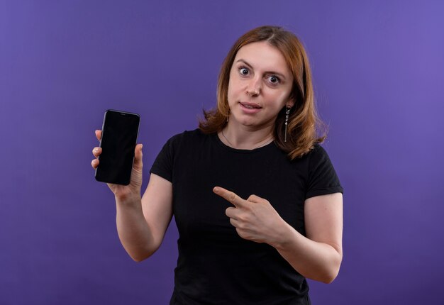 Удивленная молодая случайная женщина держит мобильный телефон и указывает на него на изолированной фиолетовой стене