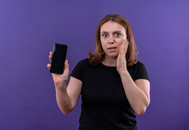 Удивленная молодая случайная женщина держит мобильный телефон и кладет руку на щеку на изолированной фиолетовой стене с копией пространства