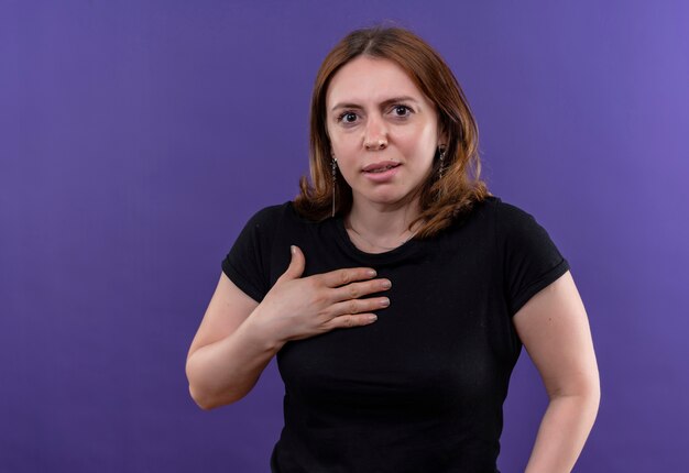 Удивленная молодая случайная женщина кладет руку на грудь на изолированной фиолетовой стене с копией пространства