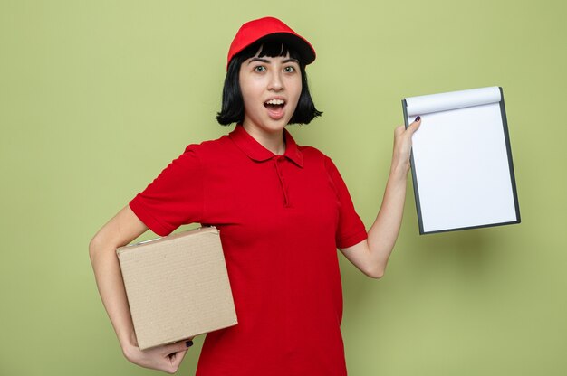 Surprised young caucasian delivery girl holding cardboard box and clipboard