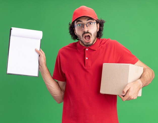 Surprised young caucasian delivery man in red uniform and cap wearing glasses holding cardbox and showing clipboard 