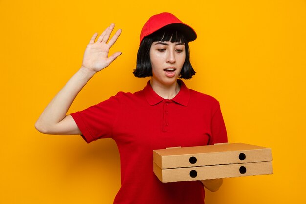 Surprised young caucasian delivery woman holding and looking at pizza boxes standing with raised hand 