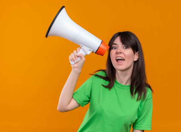 Surprised young caucasian girl in green shirt holds loud speaker and looks at camera on isolated orange background