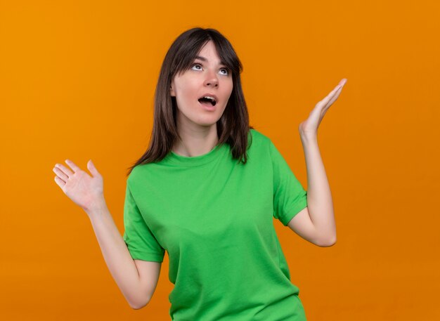 Surprised young caucasian girl in green shirt raises hands up and looks up on isolated orange background