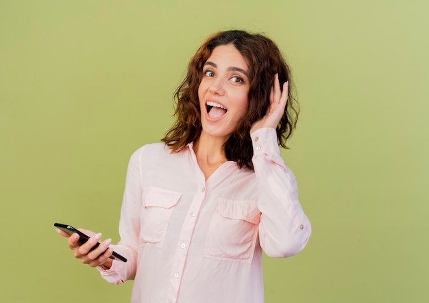 Surprised young caucasian girl holds hand behind ear trying to hear and holds phone isolated on green background with copy space