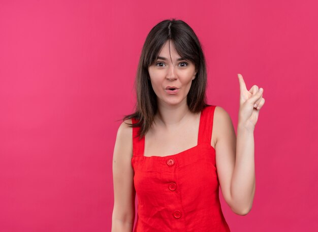 Surprised young caucasian girl pointing up and looking at camera on isolated pink background with copy space