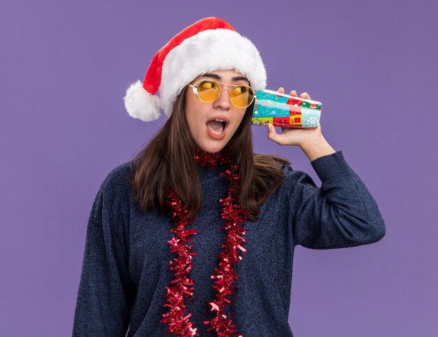 Surprised young caucasian girl in sun glasses with santa hat and garland around neck holds paper cup close to ear and looks at side isolated on purple wall with copy space