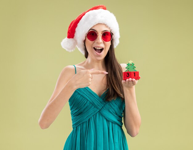 クリスマスツリーの飾りを持って指さしているサンタの帽子とサングラスで驚いた若い白人の女の子